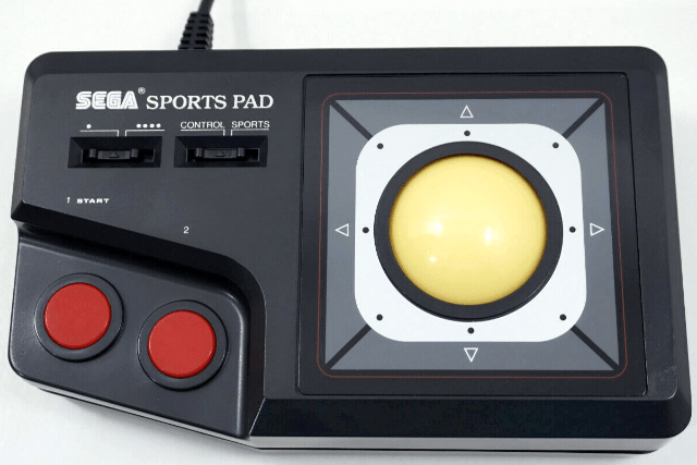 Fotografía del SEGA Sport Pad. Es negro, con una gran bola de color marfil en la parte derecha. En la parte superior izquierda hay dos pequeñas palancas. En la inferior izquierda dos botones rojos.